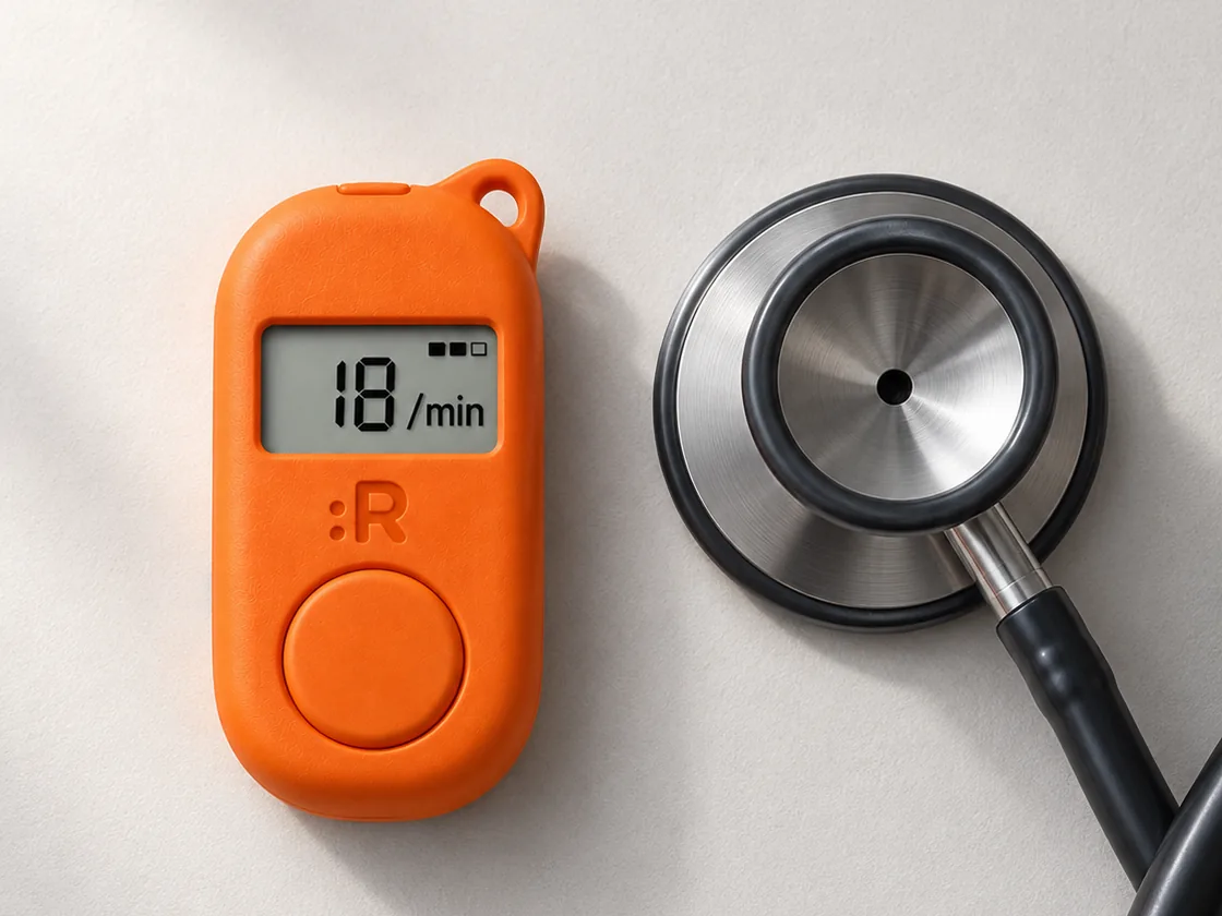 Concept render of the orange device lying beside a stethoscope chestpiece on a pale surface