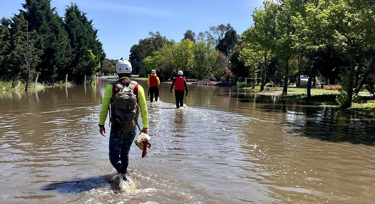 Three rescuers in helmets wading down a flooded street, seen from behind